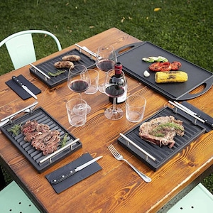Può includere: Un tavolo di legno apparecchiato per un pasto all'aperto con cibo alla griglia, vino e bicchieri. Il tavolo presenta griglie nere con bistecche e verdure cotte, una bottiglia di vino rosso e diversi calici. Sono presenti anche una forchetta e coltelli.
