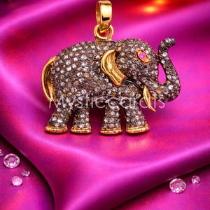 May include: A gold-toned elephant pendant encrusted with small, sparkling stones, set against a vibrant pink satin background. The elephant has a gold trunk, ears, and feet, with a small pink gem for an eye. The text "Mysticcards" is visible.