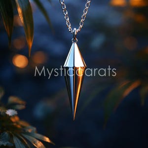 May include: A silver pendant necklace featuring a geometric, cone-shaped pendant suspended from a fine chain. The background is a blurred view of leaves and soft lighting. The text "Mystic Carats" is visible.