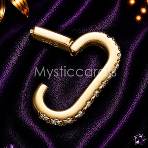 May include: A gold-colored carabiner-style clasp with a row of small, clear gemstones along one side. The clasp is marked "14K" and is set against a dark purple, textured background. The text "Mysticcarfts" is visible.