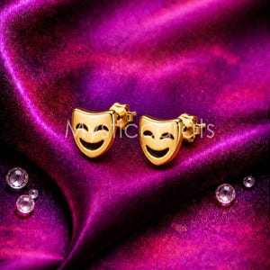 May include: A pair of gold-colored stud earrings shaped like smiling theater masks. The earrings have a black curved line for the mouth and are set against a purple satin background. The earrings are small, likely less than 1 inch in size.