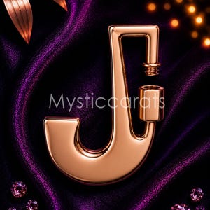 Pode incluir: Uma letra "J" dourada com um design decorativo, sobre um fundo de cetim roxo escuro. A imagem inclui o texto "Mysticcarats" e várias pedras preciosas rosa. A estética geral é luxuosa e elegante.