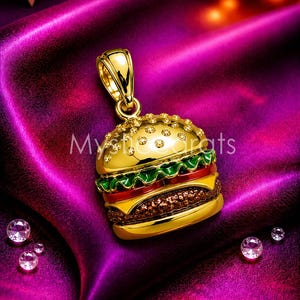 May include: A gold-colored pendant in the shape of a hamburger, with a bun, lettuce, tomato, cheese, and a patty. The pendant has a loop for a chain and is embellished with small, clear gemstones. The background is a deep purple fabric.