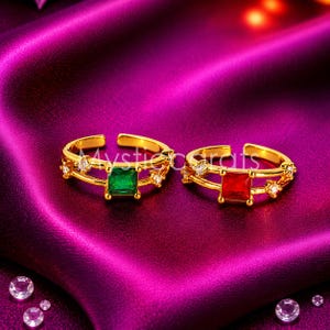 May include: Two gold-toned open rings featuring a square-cut gemstone at the centre. One ring has a vibrant green stone, while the other has a bright red stone. Both bands are accented with small, clear, round-cut stones along the sides.