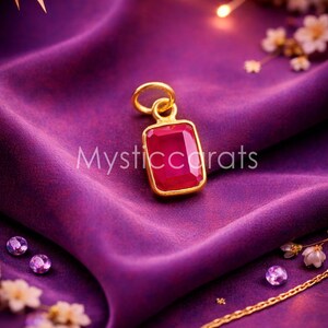 May include: A gold pendant with a rectangular, ruby-colored gemstone, set against a purple satin background. The pendant is inscribed with "Mysticcarats". Additional small gemstones and a gold chain are also visible.