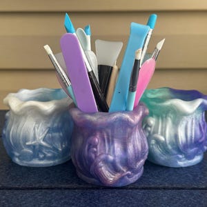 May include: Three small, round, wavy-rimmed containers with a pearlescent finish in blue, purple, and teal. The center purple container is filled with various crafting tools, including brushes, silicone sculpting tips, and plastic spatulas.