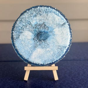 May include: A circular resin coaster featuring a swirling pattern of deep blue, light blue, and white pigments with a textured, cracked surface effect. The coaster is displayed upright on a small, light-colored wooden tripod easel stand.