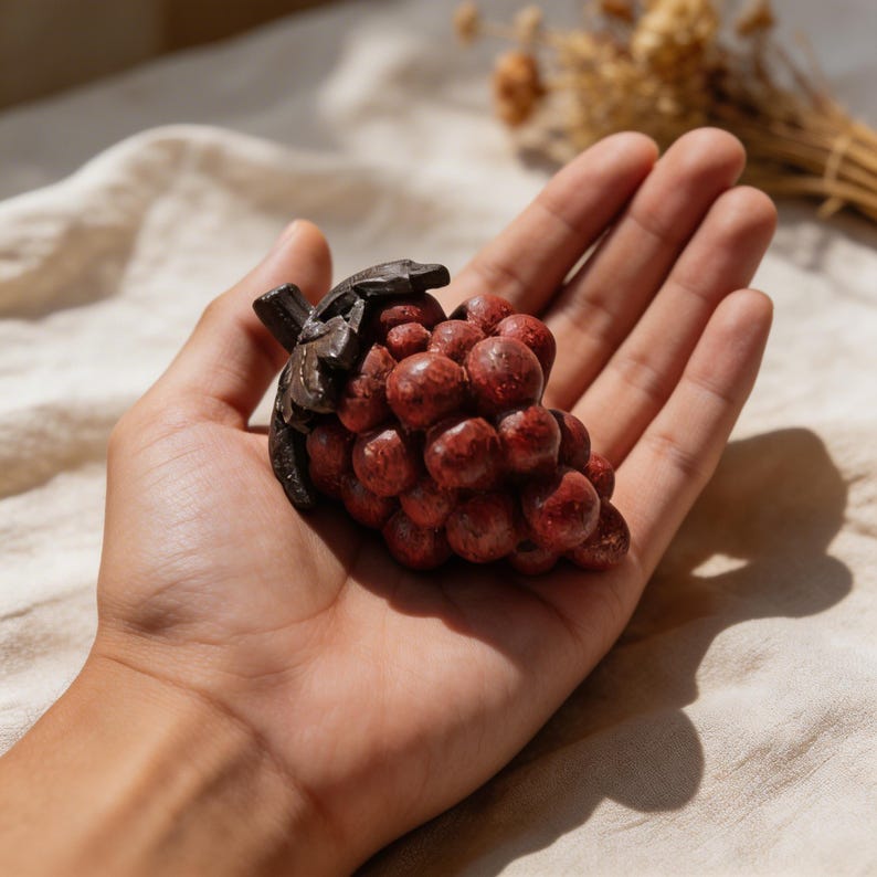 May include: A cluster of red grapes with a dark brown stem and leaves. The grapes are small and round, with a textured surface. The sculpture is held in a hand.