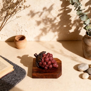 May include: A decorative cluster of red grapes rests on a small, square wooden base. The grapes are a deep burgundy color, and the wooden base has a rich, dark brown hue. The image is set against a neutral background with natural light.