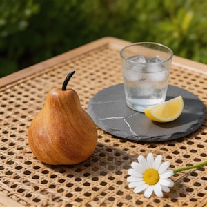 May include: A smooth, polished wooden pear figurine with a dark stem, a clear glass of water with ice cubes, a yellow lemon wedge on a round slate coaster, and a single white daisy with a yellow center on a woven cane surface.