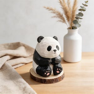 Op de afbeelding: Een gesneden houten panda beeldje met zwart-witte kleuren, zittend op een klein houten schijfje. De panda heeft zwarte oren, ogen en ledematen, met roze voetzolen. Een witte vaas met gedroogde planten staat op de achtergrond.