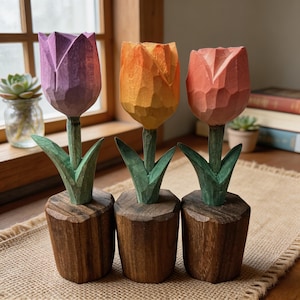 May include: Three hand-carved wooden tulips in a row. The flowers are purple, orange, and pink, with green stems and leaves. Each tulip sits in a small, brown wooden pot. The background includes a window and a succulent plant.