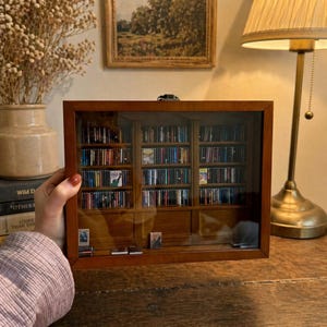 Könnte beinhalten: Ein Miniatur-Holzschrank mit Glasfront, gefüllt mit winzigen CD-Hüllen. Der Schrank hat eine warme braune Farbe und steht auf einer Holzoberfläche. Im Hintergrund befinden sich eine Lampe und ein gerahmtes Gemälde.