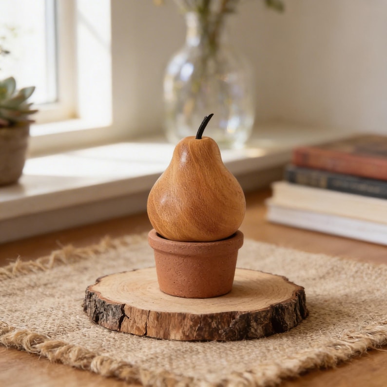 May include: A wooden pear sculpture with a black stem sits atop a small terracotta pot. The pot rests on a natural wood slice, displayed on a burlap placemat. The pear is a light brown color.