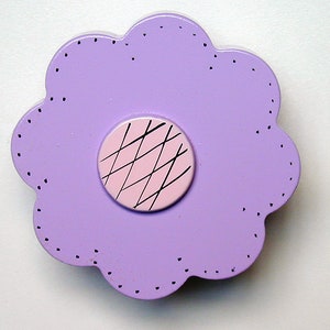 May include: A lilac-colored, flower-shaped wooden decorative item. The flower has a scalloped edge with small dotted details. The center is a pink circle with black crisscross lines.