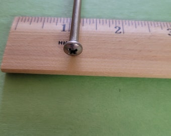 May include: A silver screw with a flat head, positioned next to a wooden ruler. The ruler shows measurements in inches. The screw is approximately 2 inches long.