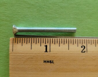 May include: A silver flat head screw with a threaded shaft, positioned next to a wooden ruler. The ruler shows measurements in inches, with the screw measuring approximately 1.5 inches in length. The background is a solid green.