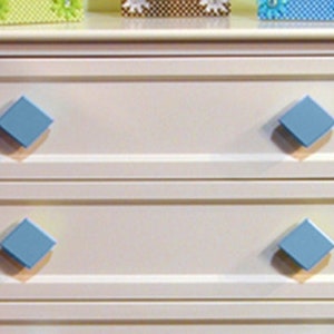 May include: White dresser with four drawers and blue diamond-shaped knobs. The dresser has a simple design and is made of wood.