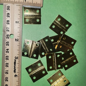May include: A close-up of ten small, gold-colored hinges. The hinges are arranged in a random pattern on a green surface. A wooden ruler with inch markings is visible in the background.