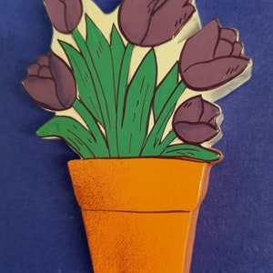 May include: A hand-painted wooden cutout of a potted plant with purple tulips and green leaves. The pot is orange.