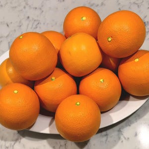 May include: A collection of bright orange citrus fruits piled high on a white oval platter. Each round fruit features a textured, dimpled peel and a small, light-colored circular stem mark at the top, creating a vibrant display of fresh produce.