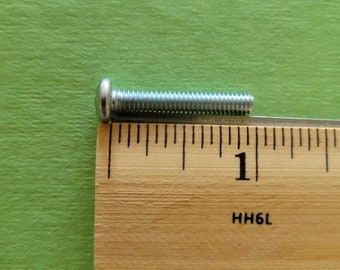 May include: A silver screw with a round head is placed next to a wooden ruler. The screw is about 1 inch long. The ruler has markings in inches and the number "1" is visible.