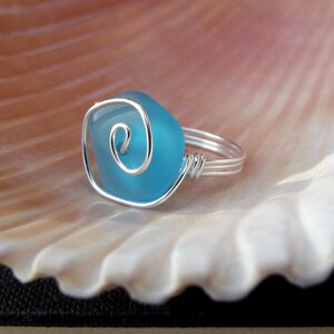 May include: A silver wire ring with a turquoise blue glass stone. The stone has a silver wire swirl design.