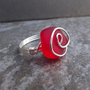 May include: A silver wire ring with a red glass square shaped stone. The wire is wrapped around the stone in a swirl design.