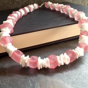 May include: A pink and white beaded necklace made of square and irregular shaped beads. The necklace is on a dark surface.