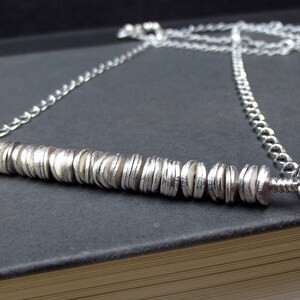May include: A silver chain necklace with a long, silver bar pendant made of stacked, overlapping discs.