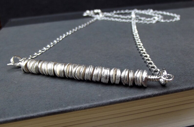 May include: A silver chain necklace with a long, silver bar pendant made of stacked, overlapping discs.