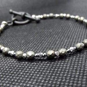 May include: A silver and gray beaded bracelet with a black metal clasp. The beads are faceted and have a rough, natural texture.
