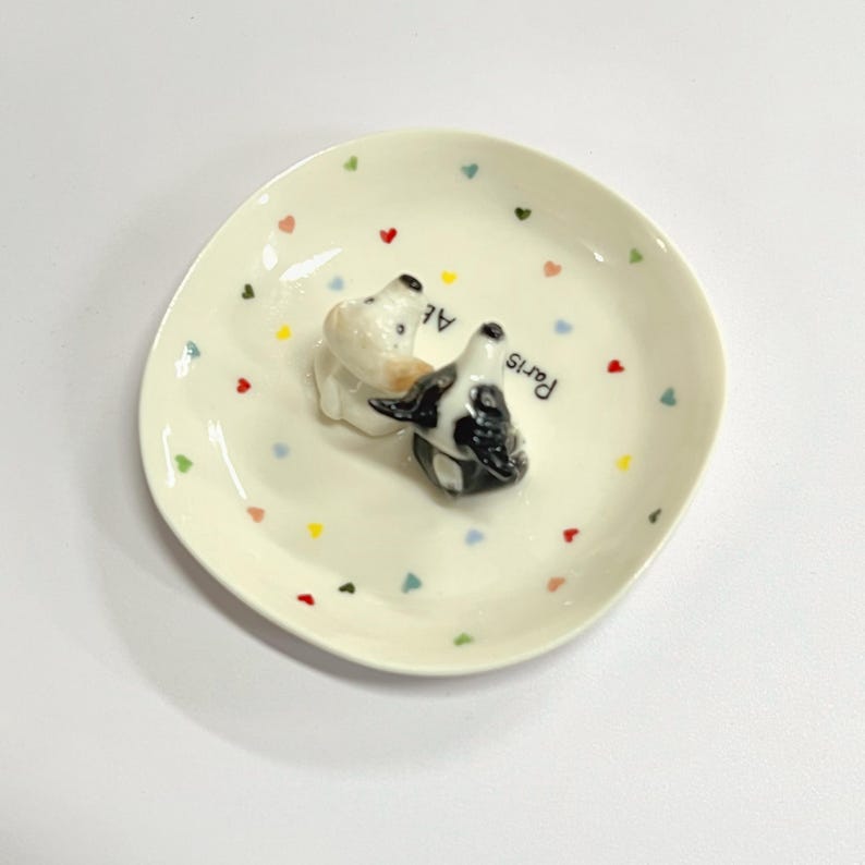 May include: A small, off-white ceramic plate with a slightly irregular shape. The plate is decorated with colorful heart-shaped accents and features a 3D sculpture of two dogs. The word "Paris" is written on the side.