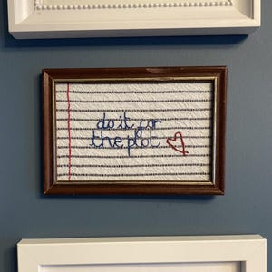 May include: A rectangular dark brown wooden frame containing white fabric embroidered to resemble lined notebook paper. The embroidery features the blue text "do it for the plot" and a small red stitched heart on the right side.