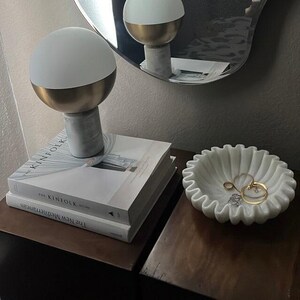 May include: A modern bedside table arrangement featuring a white and gold lamp, a stack of books, and a white marble dish holding rings. The lamp has a gold base and a white globe. The books are titled "The Kinfolk" and "The New Mediterranean".