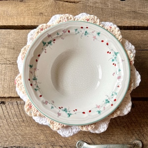 May include: A round ceramic bowl with a cream-colored base and a delicate rim pattern of green leaves, small pink flowers, and red berries. The bowl rests on a layered, scalloped-edge crochet doily featuring alternating white and beige yarn.