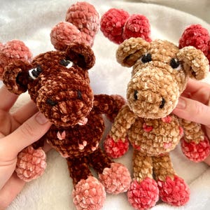 May include: Two handmade crochet moose toys. One is dark brown, the other is tan. Both have red and pink antlers and feet. The moose have black eyes and small pink hearts on their chests. The toys are soft and plush.
