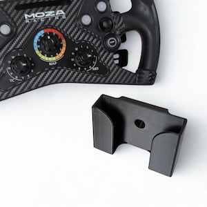 May include: A black racing wheel with the text "MOZA RACING" and a black mounting bracket. The wheel has multiple buttons and dials, and a carbon fibre pattern. The bracket is rectangular with a circular hole.