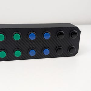 May include: A black rectangular device with a carbon fiber texture, featuring twelve round buttons. The buttons are arranged in three rows, with green, blue, and black buttons. The device is likely an electronic control panel.