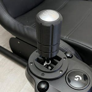 May include: Black and silver gear shift lever with a textured grip, mounted on a black base. The base has a silver button with a 'G' logo. The background shows a black seat.