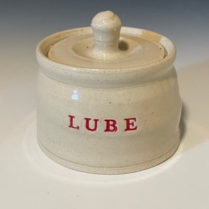 May include: A white ceramic jar with a lid and the word "LUBE" written in red lettering.