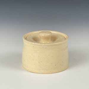 May include: A small, round, off-white ceramic jar with a matching lid. The lid has a small, rounded handle. The jar and lid have a speckled appearance, suggesting a handmade or rustic aesthetic. The jar is likely used for storage.