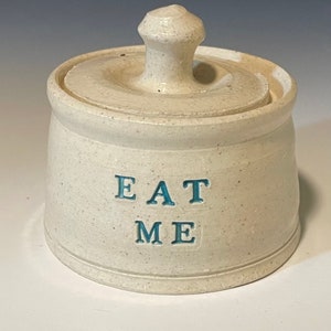 May include: White ceramic jar with a lid and a blue inscription that reads "EAT ME".
