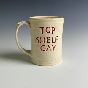 May include: A white ceramic mug with a red inscription that reads "TOP SHELF GAY".
