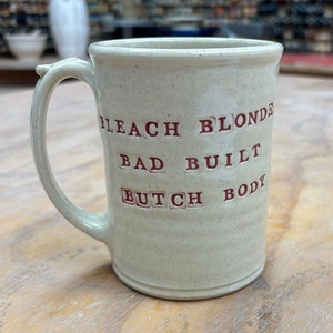 May include: A white ceramic mug with a speckled glaze. The mug has a red inscription that reads "Bleach Blonde Bad Built Butch Body".