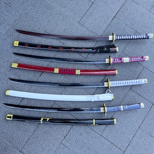 May include: An assortment of Japanese katana swords with various handle and scabbard designs. The blades are black, and the handles feature colors like red, white, and blue. Gold-colored accents are present on the hilts and scabbards.