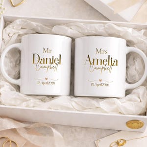 Peut inclure: Deux mugs en céramique blanche avec des inscriptions dorées, présentés dans un coffret cadeau blanc. L'un des mugs indique "Mr Daniel Campbell" et l'autre "Mrs Amelia Campbell", tous deux avec une date en dessous. La boîte est remplie de papier de soie blanc.