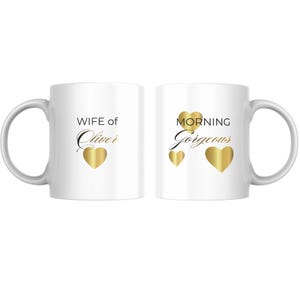Op de afbeelding: Twee witte keramische mokken. Op de ene mok staat "WIFE of Oliver" met een gouden hart. Op de andere mok staat "MORNING Gorgeous" met drie gouden harten. Beide mokken hebben een witte handgreep.
