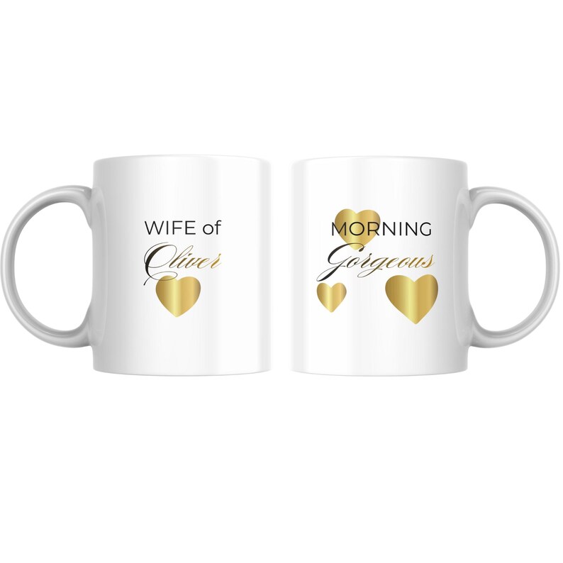 Op de afbeelding: Twee witte keramische mokken. Op de ene mok staat "WIFE of Oliver" met een gouden hart. Op de andere mok staat "MORNING Gorgeous" met drie gouden harten. Beide mokken hebben een witte handgreep.