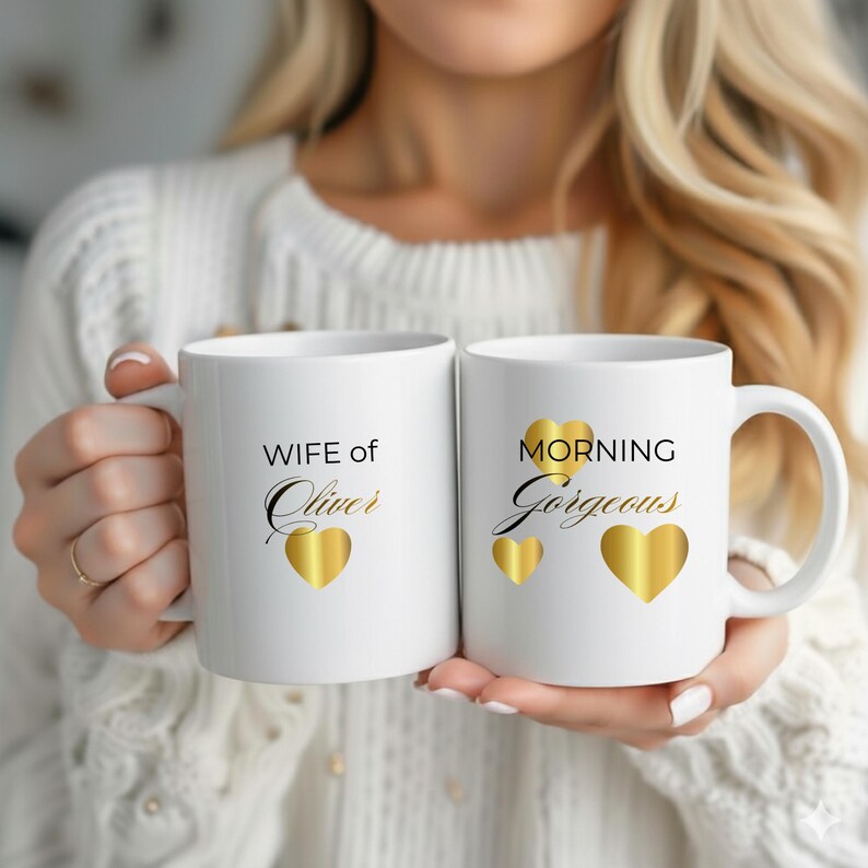 Op de afbeelding: Twee witte keramische mokken met gouden hartaccenten. E&eacute;n mok leest "WIFE of Oliver" en de andere "MORNING Gorgeous". De mokken worden vastgehouden voor een witte trui.